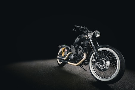 A sleek black motorcycle with a matte finish, displayed against a dark background. The bike features whitewall tires, chrome accents, and a low seat design, creating a classic yet modern look. The headlight is prominently positioned, and the handlebar is low, enhancing its streamlined appearance.