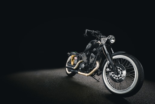 A sleek black motorcycle with a matte finish, displayed against a dark background. The bike features whitewall tires, chrome accents, and a low seat design, creating a classic yet modern look. The headlight is prominently positioned, and the handlebar is low, enhancing its streamlined appearance.