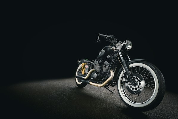 A sleek black motorcycle with a matte finish, displayed against a dark background. The bike features whitewall tires, chrome accents, and a low seat design, creating a classic yet modern look. The headlight is prominently positioned, and the handlebar is low, enhancing its streamlined appearance.