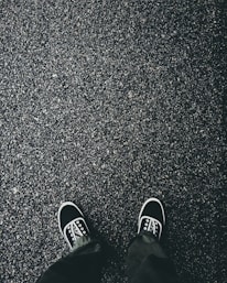 A pair of black sneakers with white soles and laces are visible on a textured asphalt surface. The ground appears wet, suggesting it might have recently rained. The shoes are positioned as if the person wearing them is looking down.