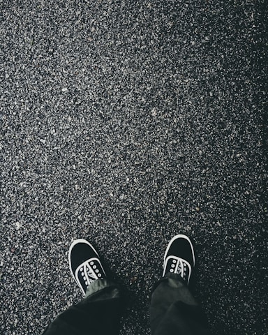 A pair of black sneakers with white soles and laces are visible on a textured asphalt surface. The ground appears wet, suggesting it might have recently rained. The shoes are positioned as if the person wearing them is looking down.