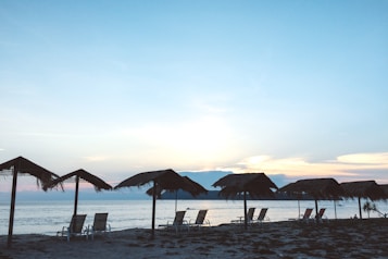 A serene beach scene with several lounge chairs arranged under thatched umbrellas facing the ocean. The sun is setting or rising, casting a gentle glow across the sky which transitions from soft pinks to light blues. The tranquil water reflects the colors of the sky, creating a peaceful atmosphere.