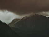 Dark, ominous clouds hover over a rugged mountain landscape, casting deep shadows over the rocky terrain. The atmosphere is moody and atmospheric, with a foreboding sense of an impending storm.