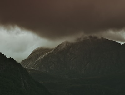 Dark, ominous clouds hover over a rugged mountain landscape, casting deep shadows over the rocky terrain. The atmosphere is moody and atmospheric, with a foreboding sense of an impending storm.