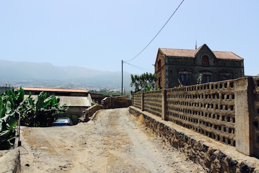 A rural dirt road leads past an old, weathered building with a tile roof. To the left, banana plants grow near a small structure with a corrugated metal roof. In the background, hazy mountains stretch under a clear blue sky.