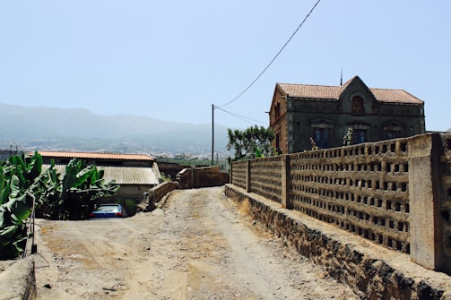 A rural dirt road leads past an old, weathered building with a tile roof. To the left, banana plants grow near a small structure with a corrugated metal roof. In the background, hazy mountains stretch under a clear blue sky.