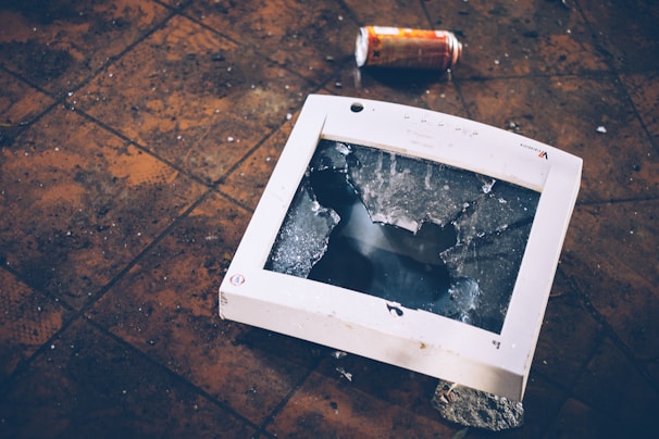 A broken computer monitor lies on a tiled floor with a shattered screen. A crumpled can is visible in the background, adding to the sense of disarray. The floor tiles appear dirty and worn, contributing to a neglected atmosphere.