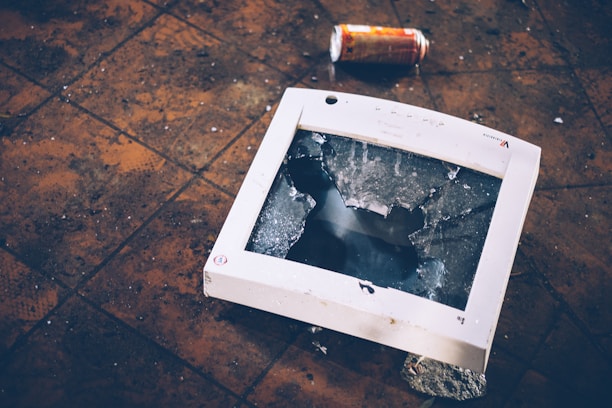 A broken computer monitor lies on a tiled floor with a shattered screen. A crumpled can is visible in the background, adding to the sense of disarray. The floor tiles appear dirty and worn, contributing to a neglected atmosphere.