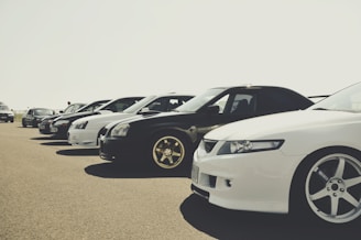 A lineup of several parked cars showcased on a clear day, emphasizing their varied models and sleek designs. The cars are positioned in a neat row, with attention to their stylish rims and lighting.