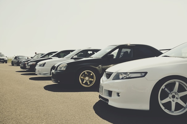 A lineup of several parked cars showcased on a clear day, emphasizing their varied models and sleek designs. The cars are positioned in a neat row, with attention to their stylish rims and lighting.