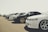 A lineup of several parked cars showcased on a clear day, emphasizing their varied models and sleek designs. The cars are positioned in a neat row, with attention to their stylish rims and lighting.