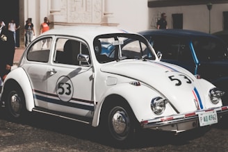 A classic white Volkswagen Beetle is parked on a street. The car features racing stripes in red and blue with the number 53 on its sides and front. Several people are visible in the background standing near a building with ornate architectural details.