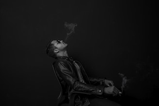 A man in a leather jacket, wearing sunglasses, is sitting on a chair with a relaxed posture. He is exhaling smoke upwards, creating a smoky trail in the air. The image has a moody and introspective feel, emphasizing the contrast between the dark tones of the background and the lighter elements.