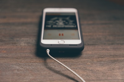 A smartphone is lying on a wooden surface with an audio cable plugged into it. The phone screen displays a blurred image, possibly of a music app, indicating media playback.