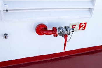 A red pipe with a valve is attached to a white industrial wall. The valve is connected to a silver metallic fitting, and a red handle is visible, likely used for controlling flow. Next to the pipe, there is a safety or identifier sign marked with 'F2'. A smaller device is mounted on the wall near the setup.