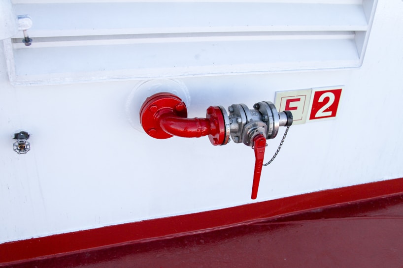 A red pipe with a valve is attached to a white industrial wall. The valve is connected to a silver metallic fitting, and a red handle is visible, likely used for controlling flow. Next to the pipe, there is a safety or identifier sign marked with 'F2'. A smaller device is mounted on the wall near the setup.