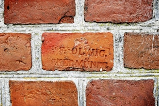 A close-up view of a brick wall with rectangular bricks laid in a horizontal pattern. One of the bricks has an inscription with two words, 'F. OLWIG' and 'CREMNDE'. The bricks are a mix of reddish-brown hues, and there are some visible textures and weathering marks.