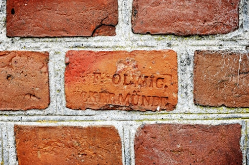 A close-up view of a brick wall with rectangular bricks laid in a horizontal pattern. One of the bricks has an inscription with two words, 'F. OLWIG' and 'CREMNDE'. The bricks are a mix of reddish-brown hues, and there are some visible textures and weathering marks.
