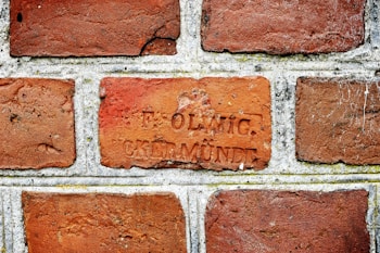 A close-up view of a brick wall with rectangular bricks laid in a horizontal pattern. One of the bricks has an inscription with two words, 'F. OLWIG' and 'CREMNDE'. The bricks are a mix of reddish-brown hues, and there are some visible textures and weathering marks.