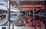 A brightly lit, expansive indoor shopping mall features a bustling food court area. Numerous eateries and dining tables are organized in the center, with people walking and sitting throughout the space. Retail stores line both levels, displaying prominent signage such as 'Westfield' and 'hmv'. The architecture includes a modern, high ceiling with warm lighting and wooden accents.
