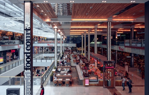 A brightly lit, expansive indoor shopping mall features a bustling food court area. Numerous eateries and dining tables are organized in the center, with people walking and sitting throughout the space. Retail stores line both levels, displaying prominent signage such as 'Westfield' and 'hmv'. The architecture includes a modern, high ceiling with warm lighting and wooden accents.