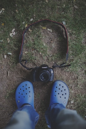 A pair of feet wearing blue clogs stand on grass, facing a digital camera placed on the ground with a strap encircling it.