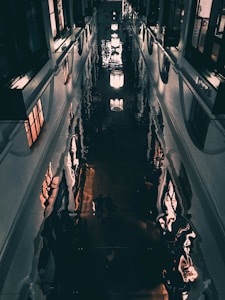 A corridor with a reflective, dark polished floor creating distorted reflections of lights and surrounding architecture, including windows and walls. The lights are hanging in a linear arrangement, casting an atmospheric glow.