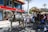 A horse-drawn carriage is situated on a city street, with a white horse pulling it. The carriage is occupied by several people, dressed in formal attire, and is passing by a building with a prominent red and white sign. The background features palm trees and a crowd of onlookers.