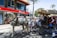 A horse-drawn carriage is situated on a city street, with a white horse pulling it. The carriage is occupied by several people, dressed in formal attire, and is passing by a building with a prominent red and white sign. The background features palm trees and a crowd of onlookers.
