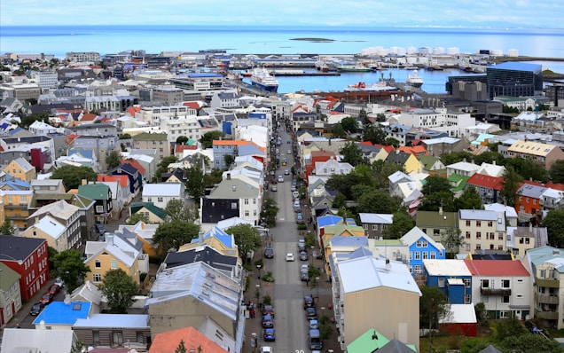 A vibrant cityscape with a variety of colorful buildings, streets lined with trees, and vehicles. In the background, a harbor with ships and industrial structures is visible, surrounded by a calm sea.