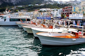 A picturesque harbor scene with several wooden boats docked along a bustling waterfront. These boats have a classic design, featuring white hulls and orange accents. People stroll along the quay, while a larger ferry is moored nearby. In the background, colorful buildings and lush hills create a scenic backdrop. Signage for tours and transportation services can be seen on the buildings.