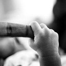 A close-up of a baby's hand gently grasping an adult's finger. The scene is captured in black and white, emphasizing the texture and softness of the baby's skin against the adult's finger. The background is blurred, keeping focus on the emotional connection depicted.
