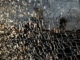 A close-up view of shattered glass forms intricate patterns, with light reflecting off the fragments creating a shimmering effect. The background is blurred, emphasizing the texture and details of the broken glass.