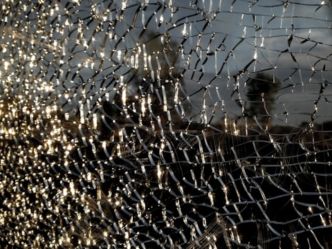 A close-up view of shattered glass forms intricate patterns, with light reflecting off the fragments creating a shimmering effect. The background is blurred, emphasizing the texture and details of the broken glass.