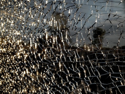 A close-up view of shattered glass forms intricate patterns, with light reflecting off the fragments creating a shimmering effect. The background is blurred, emphasizing the texture and details of the broken glass.