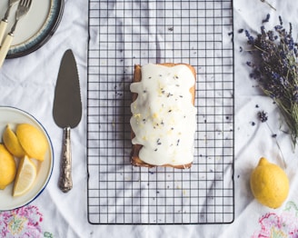 A glazed loaf cake with decorative petals is on a cooling rack, surrounded by lemons, a cake knife, and a plate with slices. A bunch of lavender is placed nearby, adding a rustic touch to the setting.