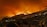 A large wildfire engulfs a hillside at night, casting an orange glow over the landscape. Smoke billows into the dark sky, and small houses are visible on the lower part of the hill, contrasting against the bright flames.