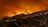 A large wildfire engulfs a hillside at night, casting an orange glow over the landscape. Smoke billows into the dark sky, and small houses are visible on the lower part of the hill, contrasting against the bright flames.