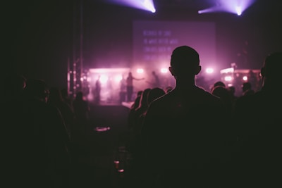 A dark auditorium with silhouetted figures standing and facing a stage where bright purple and pink lights illuminate a performance area. The atmosphere appears to be filled with a mist or haze, adding to the ambiance. A large display screen is visible at the back of the stage.