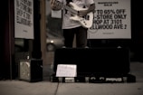 A person stands outside a store playing an electric guitar, with an amplifier to the side and a guitar case on the ground. A handwritten sign in the case reads 'Furthering Music Career.' Advertisement signs on the store window offer discounts up to 65%. The setting has a low-light, street ambiance.