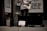 A person stands outside a store playing an electric guitar, with an amplifier to the side and a guitar case on the ground. A handwritten sign in the case reads 'Furthering Music Career.' Advertisement signs on the store window offer discounts up to 65%. The setting has a low-light, street ambiance.