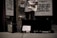 A person stands outside a store playing an electric guitar, with an amplifier to the side and a guitar case on the ground. A handwritten sign in the case reads 'Furthering Music Career.' Advertisement signs on the store window offer discounts up to 65%. The setting has a low-light, street ambiance.