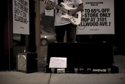 A person stands outside a store playing an electric guitar, with an amplifier to the side and a guitar case on the ground. A handwritten sign in the case reads 'Furthering Music Career.' Advertisement signs on the store window offer discounts up to 65%. The setting has a low-light, street ambiance.