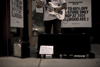 A person stands outside a store playing an electric guitar, with an amplifier to the side and a guitar case on the ground. A handwritten sign in the case reads 'Furthering Music Career.' Advertisement signs on the store window offer discounts up to 65%. The setting has a low-light, street ambiance.