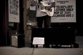 A person stands outside a store playing an electric guitar, with an amplifier to the side and a guitar case on the ground. A handwritten sign in the case reads 'Furthering Music Career.' Advertisement signs on the store window offer discounts up to 65%. The setting has a low-light, street ambiance.