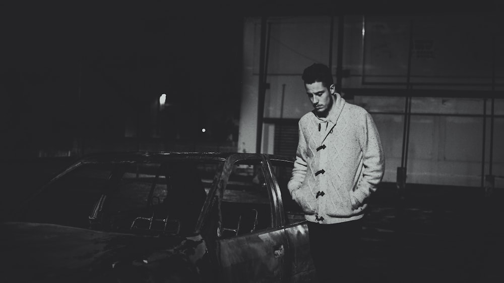 grayscale photo of man standing beside car