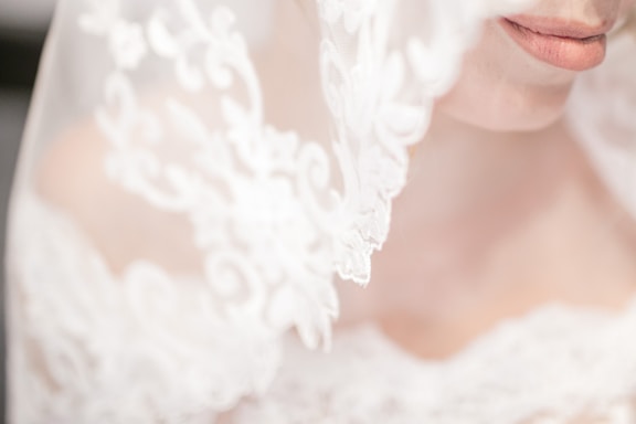 An elegant close-up of a bride's face partially obscured by a delicate, lace veil. The focus is on the lips and intricate lace patterns that are softly lit, creating a dreamy and romantic atmosphere.