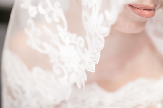 An elegant close-up of a bride's face partially obscured by a delicate, lace veil. The focus is on the lips and intricate lace patterns that are softly lit, creating a dreamy and romantic atmosphere.