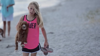 A young girl with long blonde hair is holding a small brown dog in her left hand and a pair of flip-flops in her right hand. She is wearing a bright pink shirt with the word 'LEADER' on it, along with black shorts. The background appears to be a sandy beach, with another person and another dog visible in the distance.