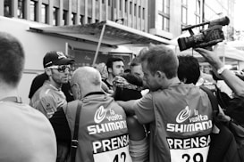A crowd of journalists gathered around a cyclist, capturing interviews and images. The cyclist is wearing sunglasses and a cap, appearing focused and calm. Some individuals in the crowd are wearing vests labeled 'la vuelta SHIMANO' and 'PRENSA', indicating they are members of the press. The scene is busy with attention focused on the cyclist, while a large video camera is also capturing the moment.